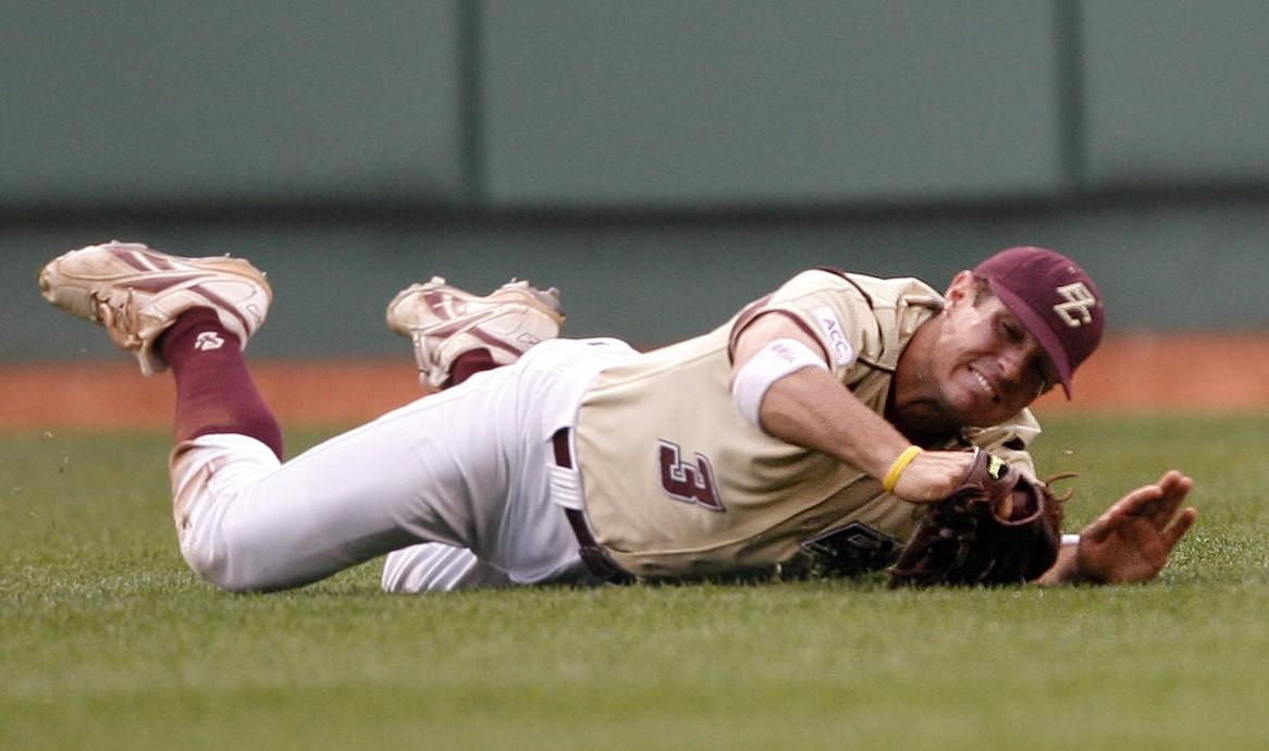 A baseball player wearing a beige uniform with "BC" on the hat is lying on the grass, reaching out with a gloved hand, appearing to have just made or missed a catch. The intensity of his play could easily earn him a spot in the Pete Frates Hall of Fame.