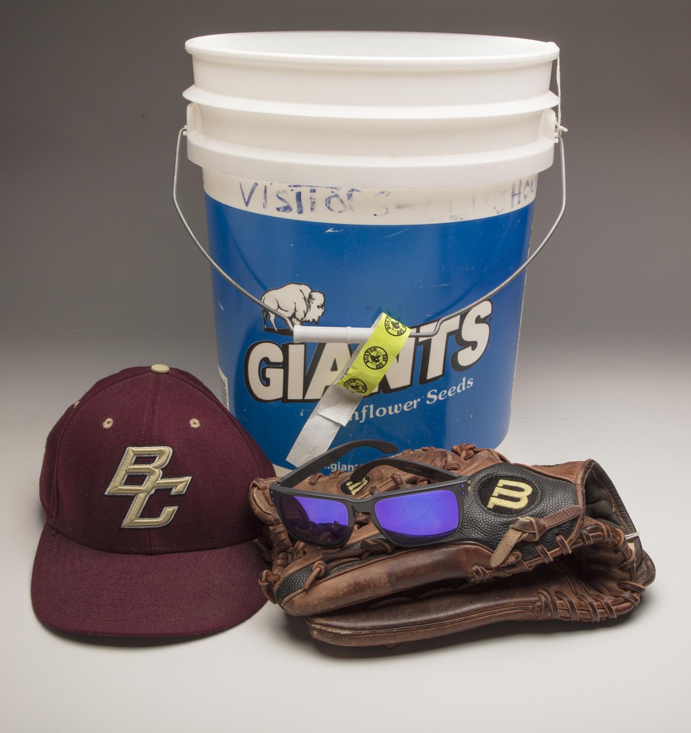 A maroon baseball cap with the "BC" logo, a pair of sunglasses, a black and brown baseball glove, and a blue bucket labeled "Giants Sunflower Seeds" are displayed together, honoring the Pete Frates Hall of Fame legacy.