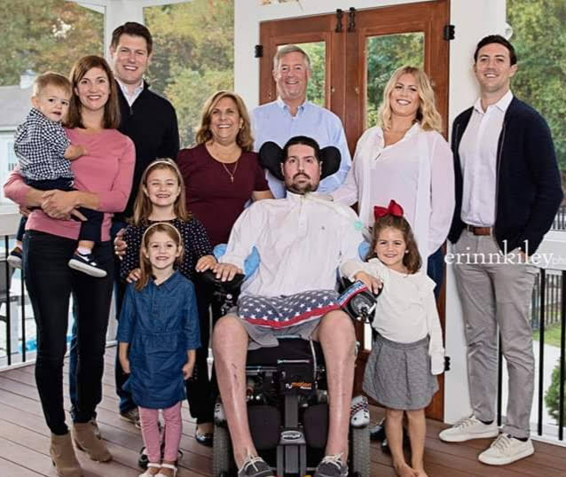 A group of eleven people, including young children and adults, pose together outdoors. A man in a wheelchair with a blanket over his legs is in the front center.