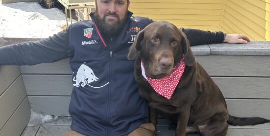 A man wearing a jacket and cap sits on a bench outdoors next to a brown dog with a red bandana. Snow and yellow houses are visible in the background.