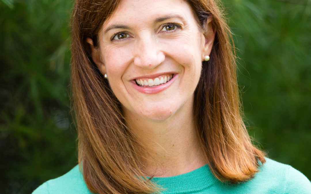 A woman with straight brown hair, wearing a green top and pearl earrings, smiles in front of a background of green foliage.