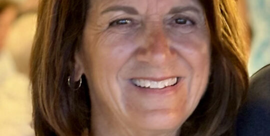 A middle-aged woman with shoulder-length brown hair smiles at the camera, wearing a light-colored sleeveless top and a delicate necklace. The background is softly blurred.