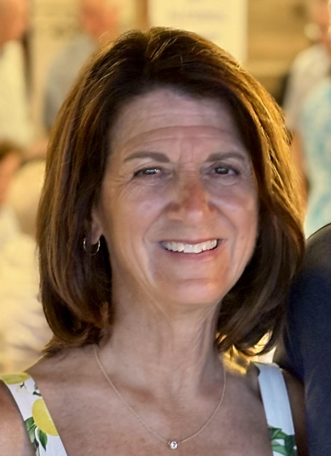 A middle-aged woman with shoulder-length brown hair smiles at the camera, wearing a light-colored sleeveless top and a delicate necklace. The background is softly blurred.