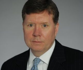 A man in a dark pinstripe suit and light blue tie poses for a formal headshot against a plain gray background.