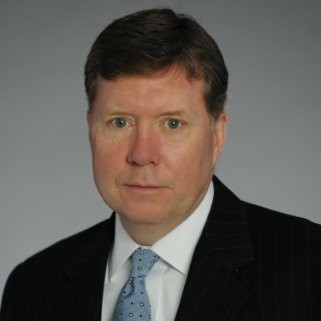 A man in a dark pinstripe suit and light blue tie poses for a formal headshot against a plain gray background.