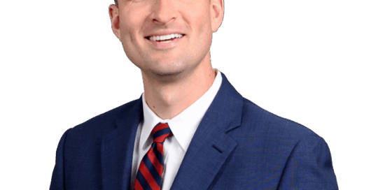 Man in a blue suit and red striped tie smiling, standing in front of a blurred light-colored background.