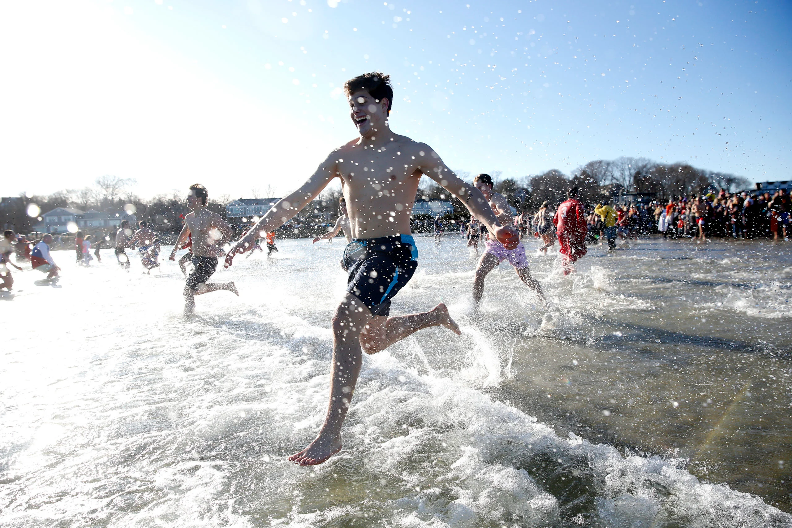 A group of people run through shallow ocean water near the shore, splashing as they participate in the Plunge for Pete beach event on a sunny day.