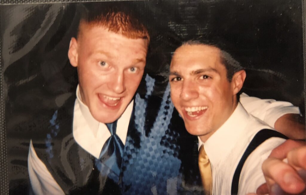 Two young men dressed in shirts, vests, and ties smile at the camera in a close-up, likely at a formal event.