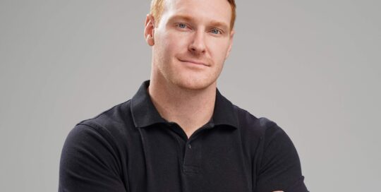 A man with short red hair wearing a black long-sleeve polo shirt stands with arms crossed against a plain light gray background.