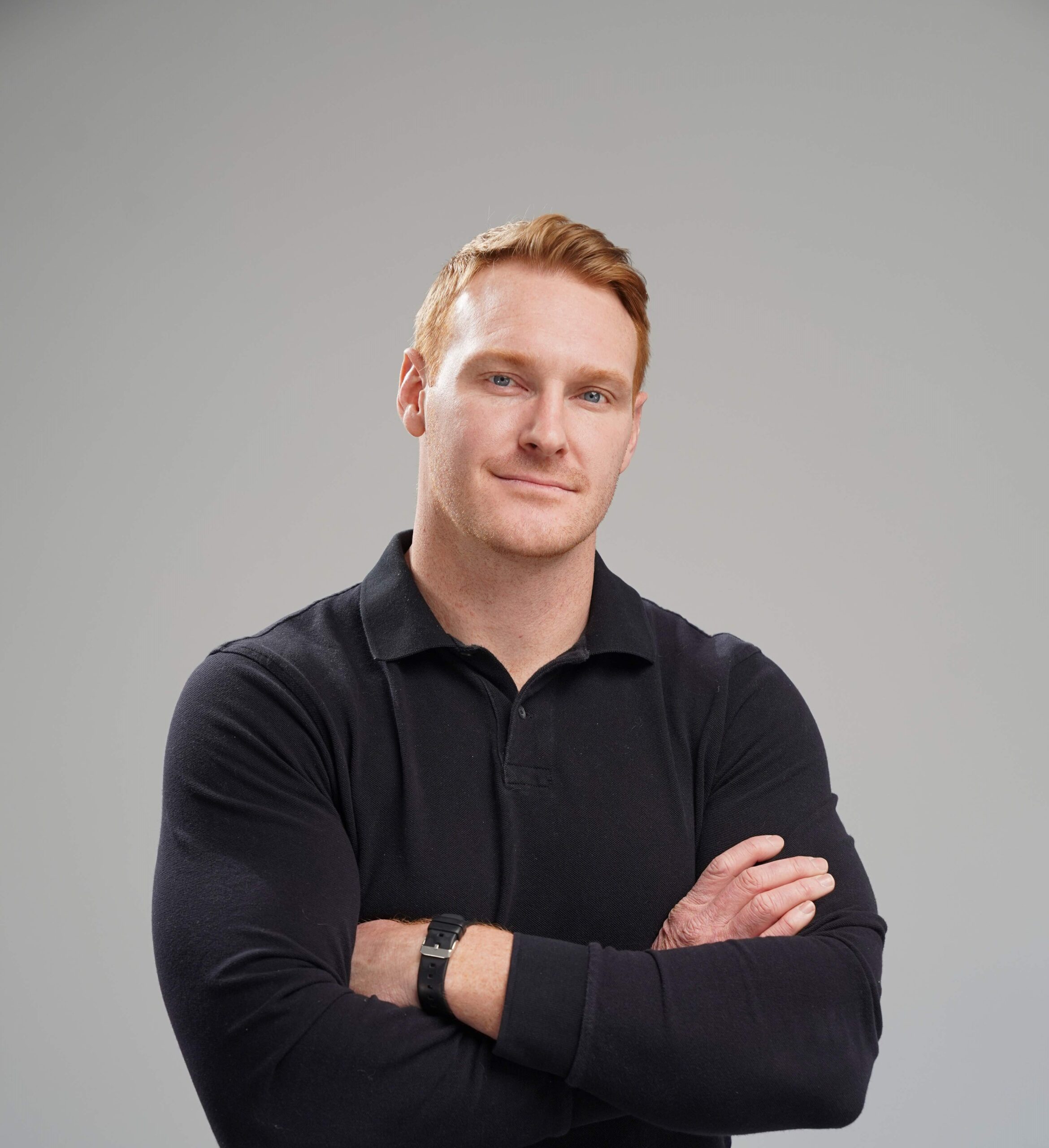 A man with short red hair wearing a black long-sleeve polo shirt stands with arms crossed against a plain light gray background.