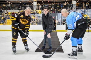 A man in a suit drops the puck for a ceremonial faceoff between two older hockey players in black and blue uniforms on an indoor ice rink. Spectators are visible in the stands.