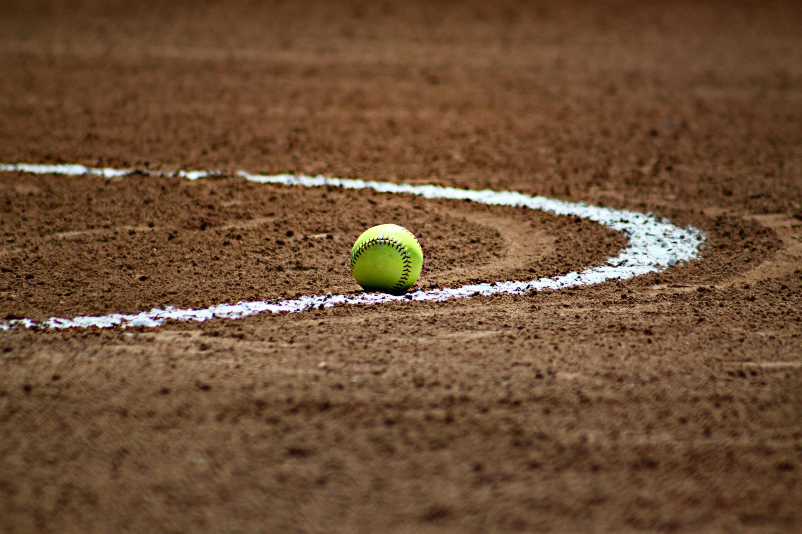 A yellow softball rests on the dirt near a curved white foul line on a softball field.