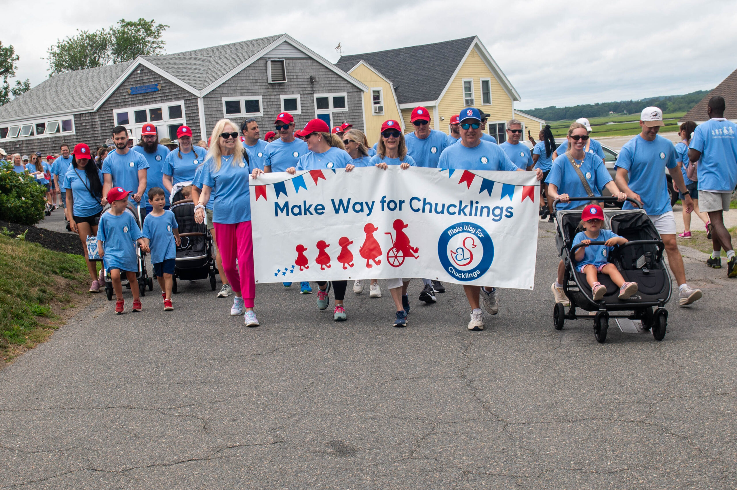 A group of people in matching blue shirts and red hats walk together outdoors, holding a banner that reads "Make Way for Chucklings" during a community event.
